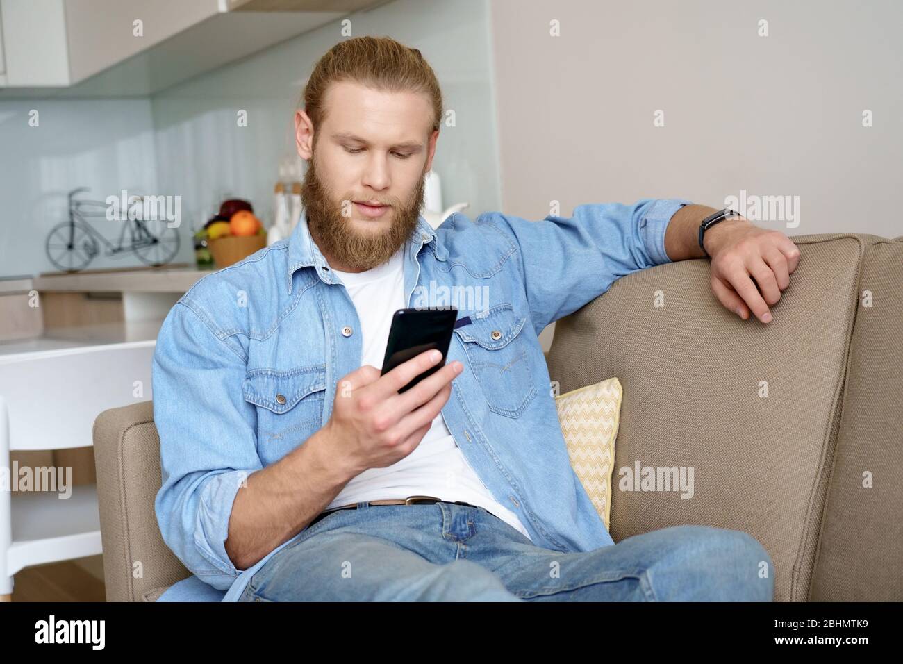 Ernst junger bärtiger Mann hält Smartphone mit Online-App auf dem Sofa zu Hause. Stockfoto