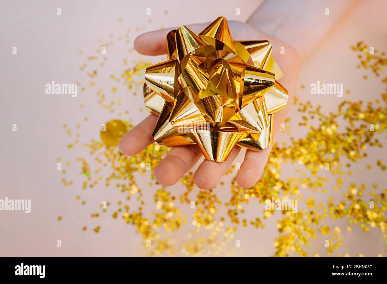 Hellhäutige Hand halten eine große Goldfolienschleife vor einem Hintergrund aus verstreuten Goldsternen auf einem weichen beigen Hintergrund. Stockfoto