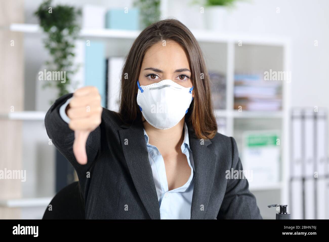 Vorderansicht einer seriösen Exekutivfrau, die eine Schutzmaske trägt und mit den Daumen nach unten zeigt und dabei die Kamera im Büro anschaut Stockfoto