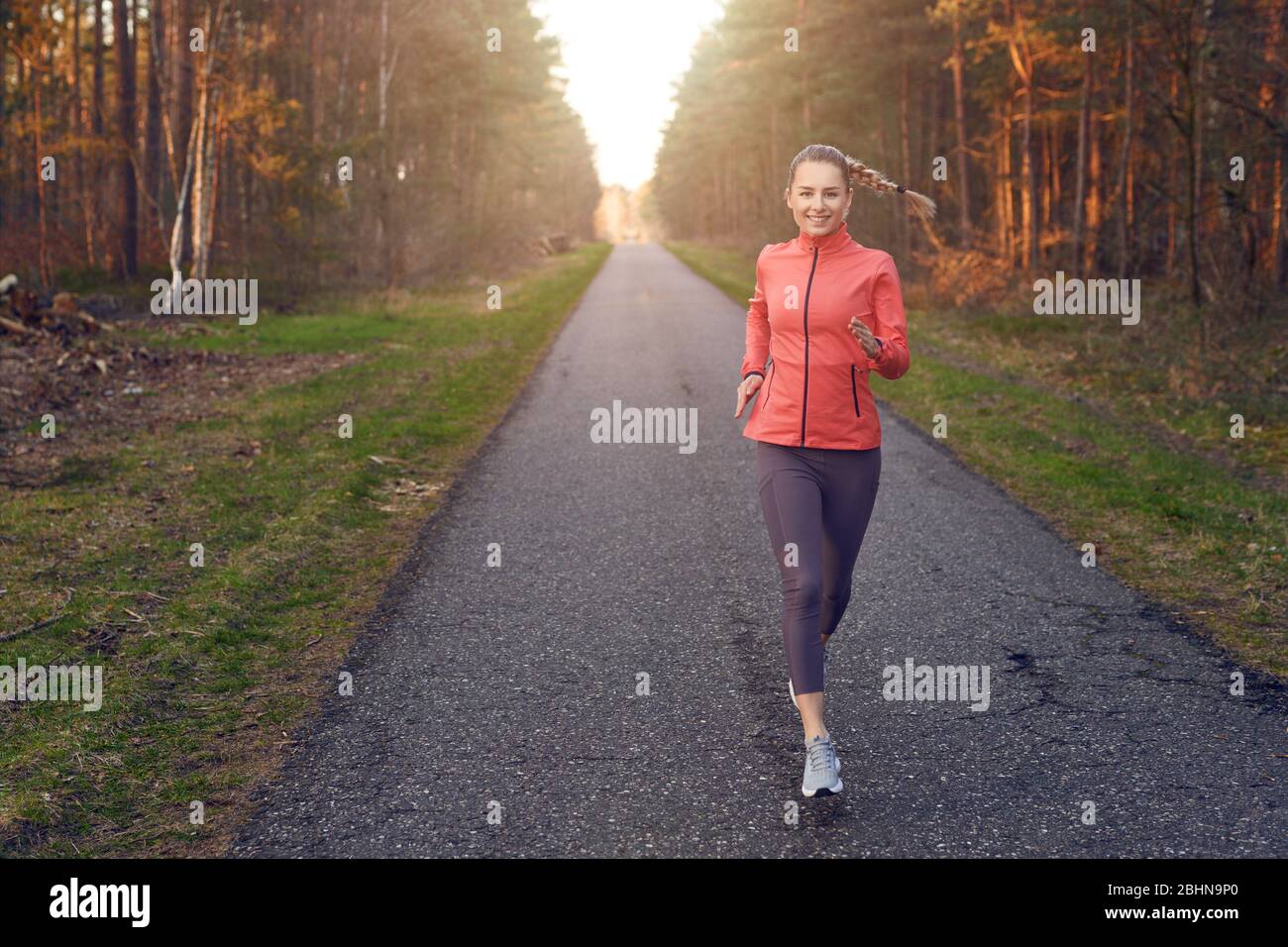 Junge flinke Frau runinng auf einer Teerspur durch den Wald im Sonnenuntergang Schein des Sonnenlichts mit Kopierraum nach links Stockfoto