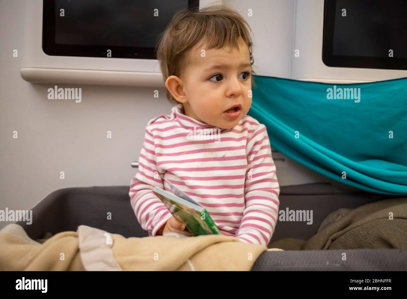 Kleines süßes Baby wachte in der Babykassette eines Flugzeugs auf, das schläfrig saß und sich umsah, Nahaufnahme, weiche Fokussierung. Stockfoto