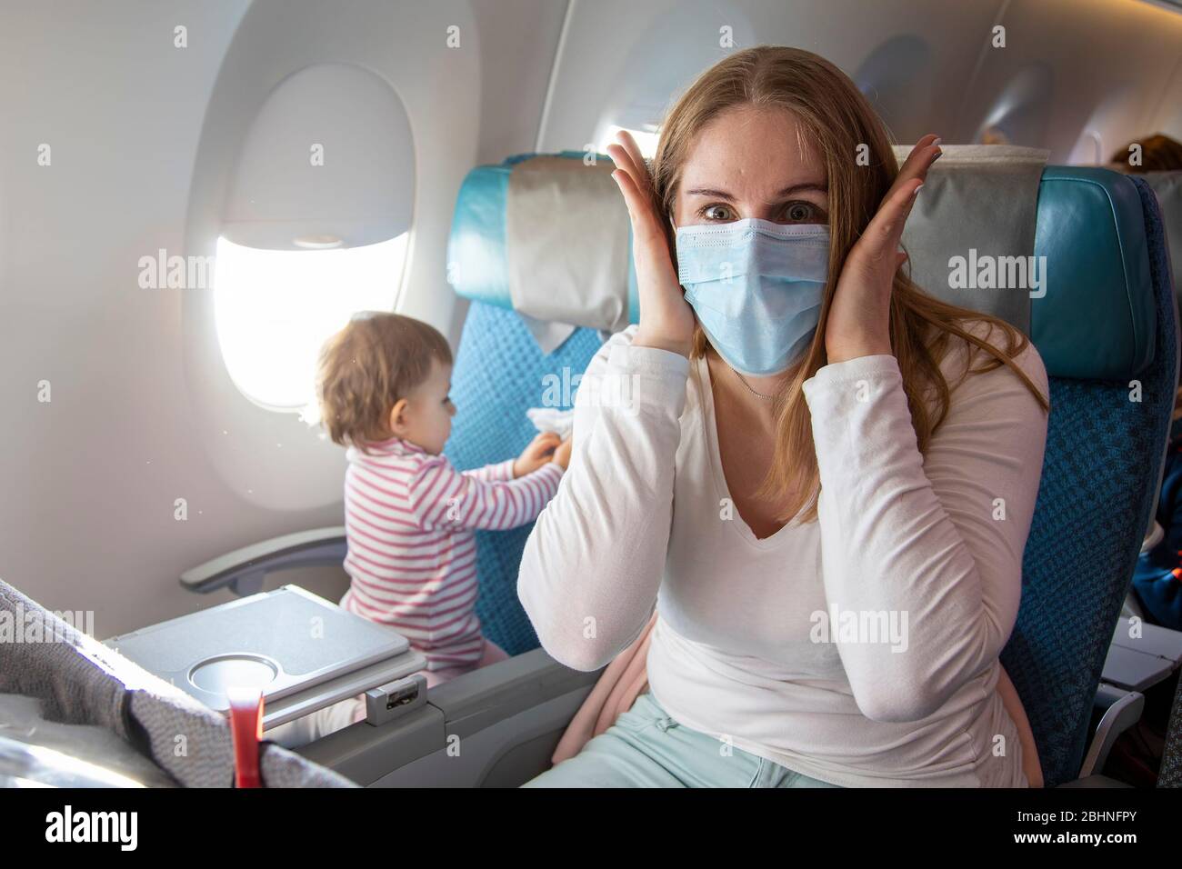Porträt einer verängstigten jungen Mutter in einem Flugzeugsessel in einer medizinischen Atemmaske, die die Kamera in Schrecken von Covid-19 Coronavirus betrachtet. A Stockfoto