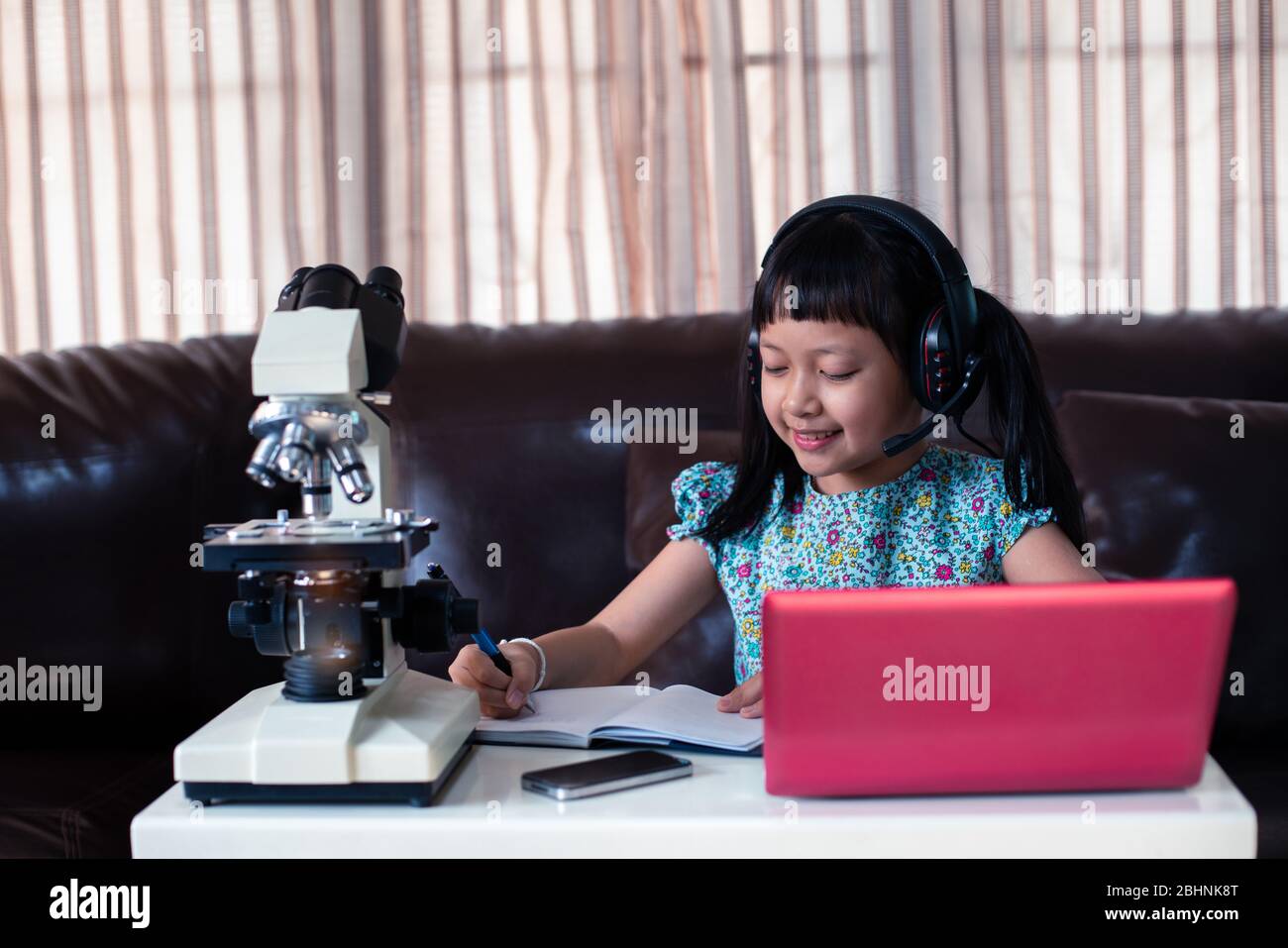 Asiatische kleines Kind Mädchen tragen Kopfhörer online lernen, indem sie Laptop und Mikroskop zu Hause, Fernunterricht Stockfoto