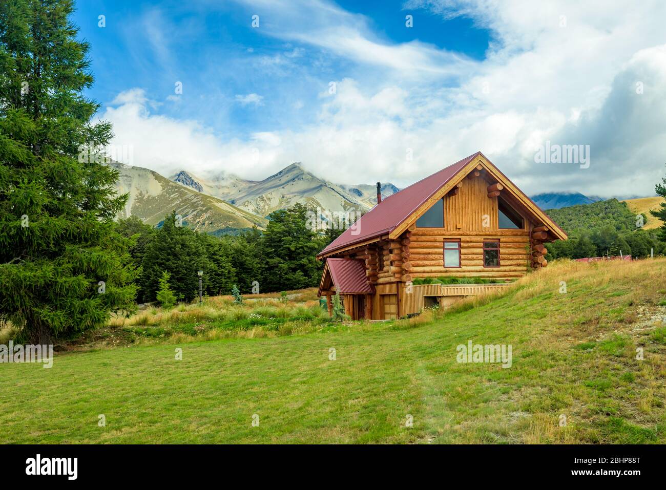 Berghütte. Log House. Solitary Cabin auf einem grünen Grasfeld mit Bäumen und einer Bergkette im Hintergrund. Stockfoto