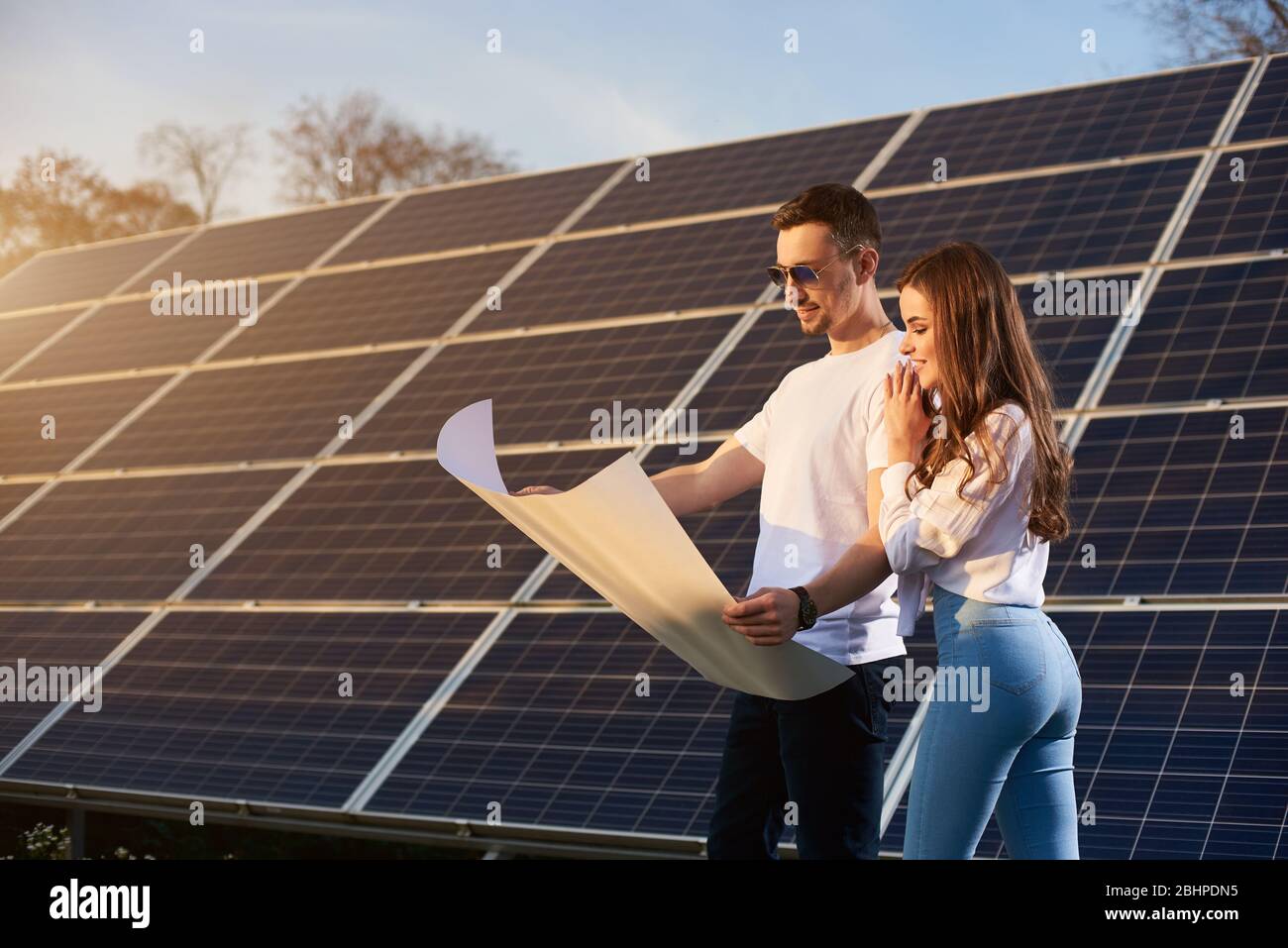Junge schöne Paar an einem sonnigen Tag steht bei einem Solarmodul und schaut in ein großes White Paper Plan, Gedanken über die Zukunft, Konzept der hellen Zukunft Stockfoto