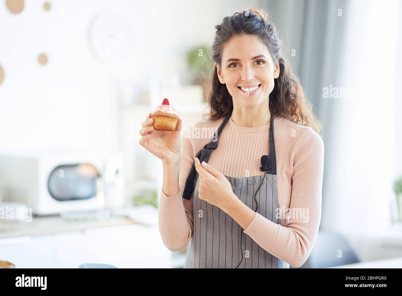 Horizontal Medium Porträt der schönen jungen Frau trägt Schürze hält hausgemachte süße Cupcake, Blick auf Kamera lächelnd Stockfoto