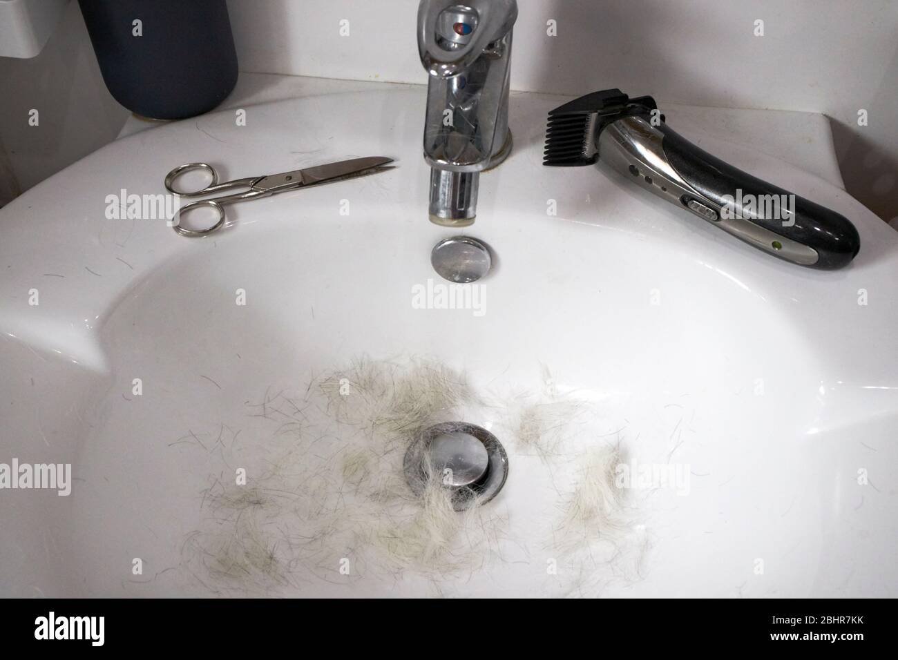 Graue Haarschere und billiger Haarschneider in einem Waschbecken aufgrund eines Mannes mittleren Alters, der seine eigenen Haare schneidet, aufgrund der Selbstisolierung Covid-19 Pandemie Stockfoto