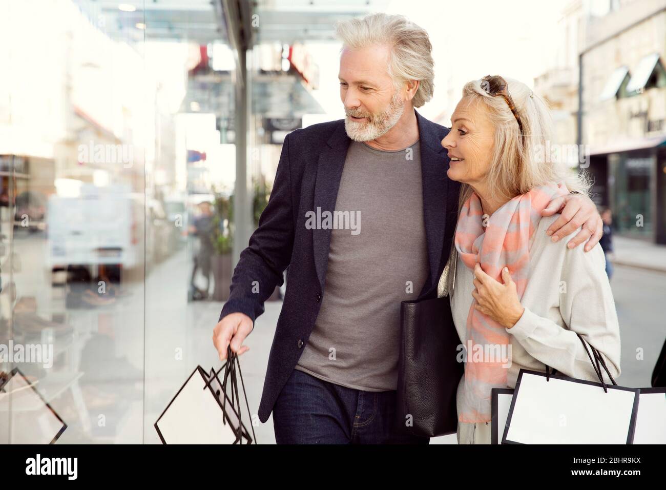 Ein Paar mit Armen um sich herum, das Einkaufstaschen trägt und in ein Schaufenster schaut. Stockfoto