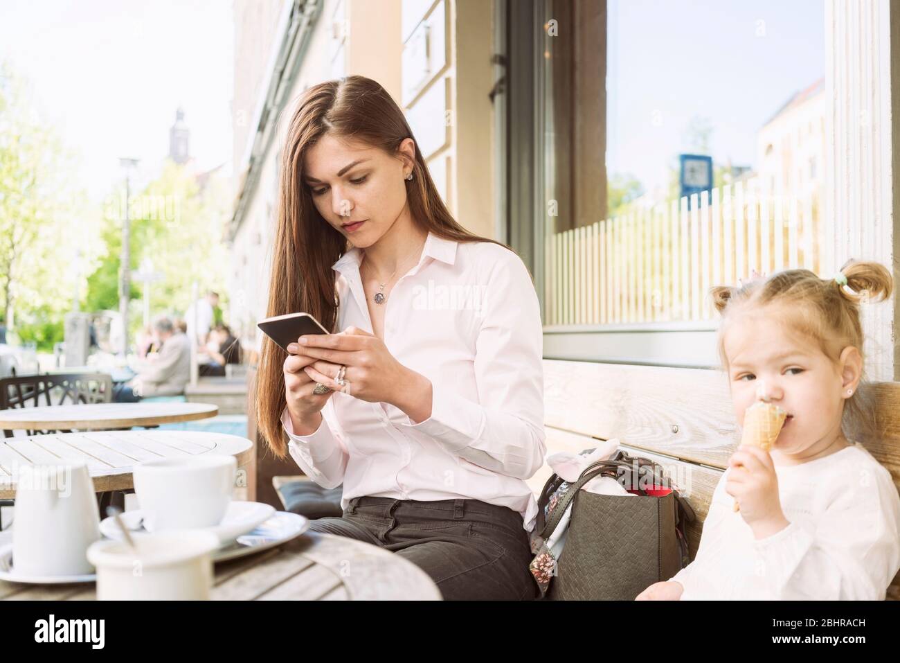 Eine Mutter und eine Tochter sitzen vor einem Café, Mutter auf ihrem Handy, Tochter essen ein Eis. Stockfoto