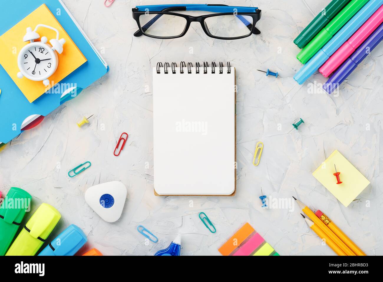 Schulpapier liegt auf einem grauen texturierten Hintergrund mit einem Notizblock in der Mitte. Die Farbe des Regenbogens, Bleistifte, Brillen, Notizblock und Wecker Stockfoto