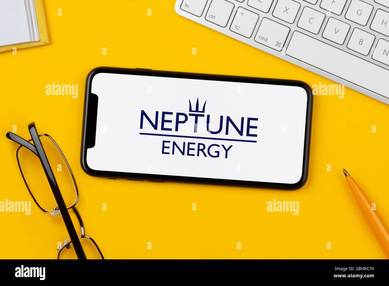 Ein Smartphone mit dem Neptune Energy Logo liegt auf einem gelben Hintergrund zusammen mit Tastatur, Brille, Stift und Buch (nur zur redaktionellen Verwendung). Stockfoto