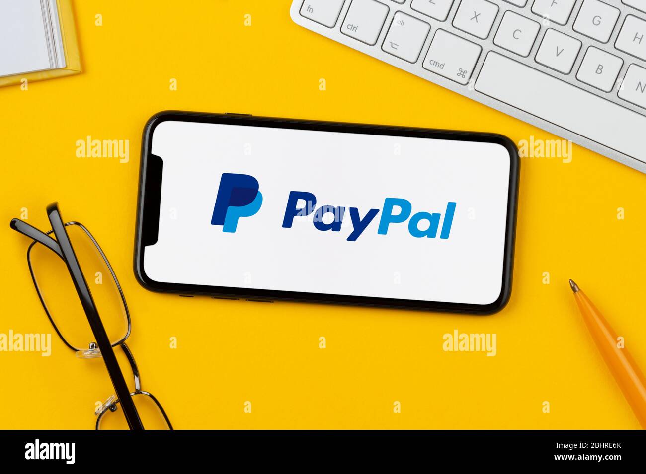 Ein Smartphone mit dem PayPal-Logo liegt auf einem gelben Hintergrund zusammen mit Tastatur, Brille, Stift und Buch (nur für redaktionelle Verwendung). Stockfoto