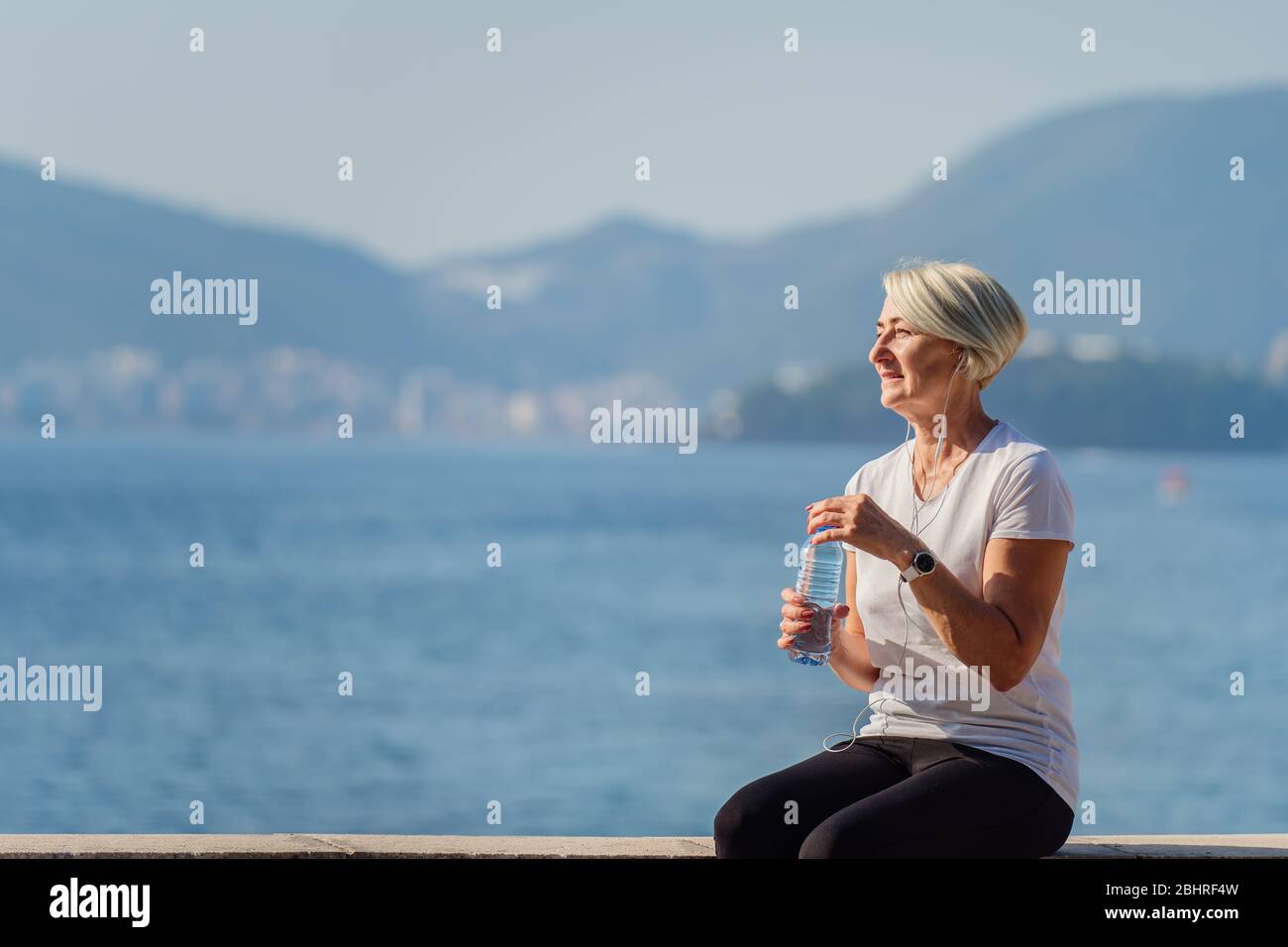 Reife Frau Trinkwasser nach dem Joggen im Hintergrund das Meer und den blauen Himmel Stockfoto