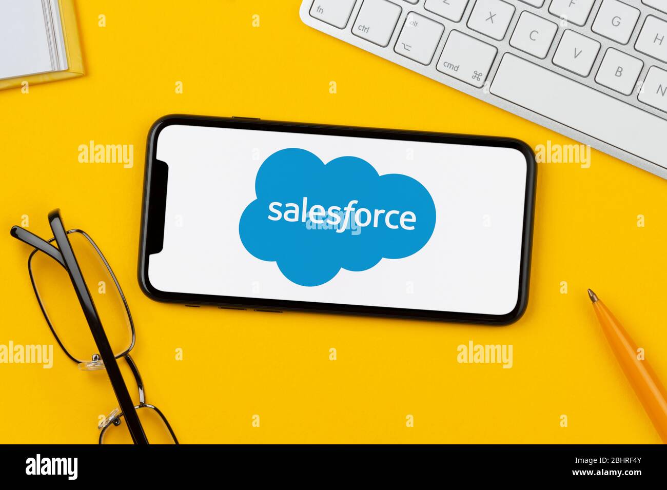 Ein Smartphone mit dem Salesforce-Logo liegt auf einem gelben Hintergrund zusammen mit Tastatur, Brille, Stift und Buch (nur redaktionelle Verwendung). Stockfoto