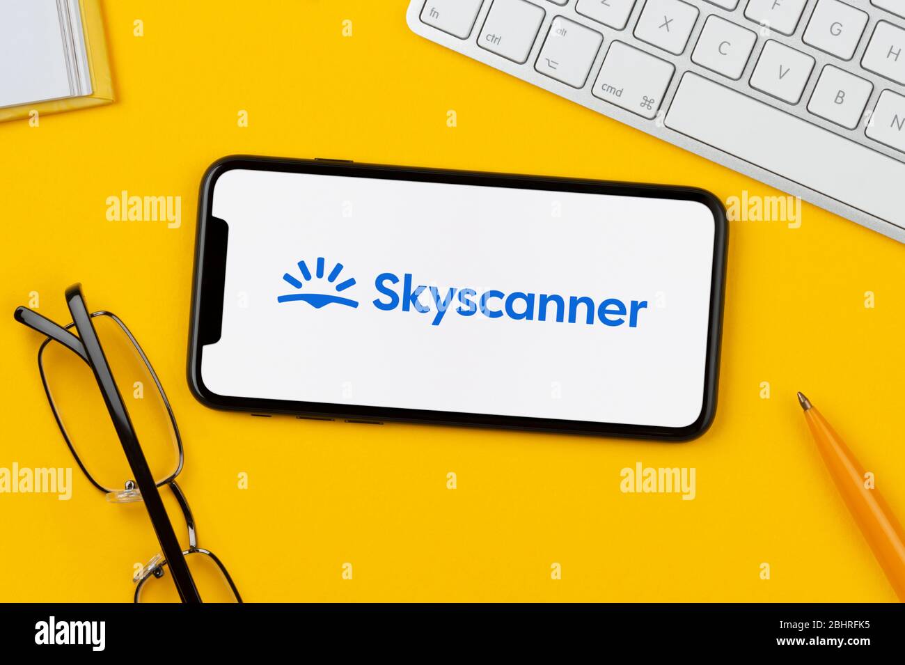 Ein Smartphone mit dem Skyscanner-Logo liegt auf einem gelben Hintergrund zusammen mit Tastatur, Brille, Stift und Buch (nur für redaktionelle Verwendung). Stockfoto