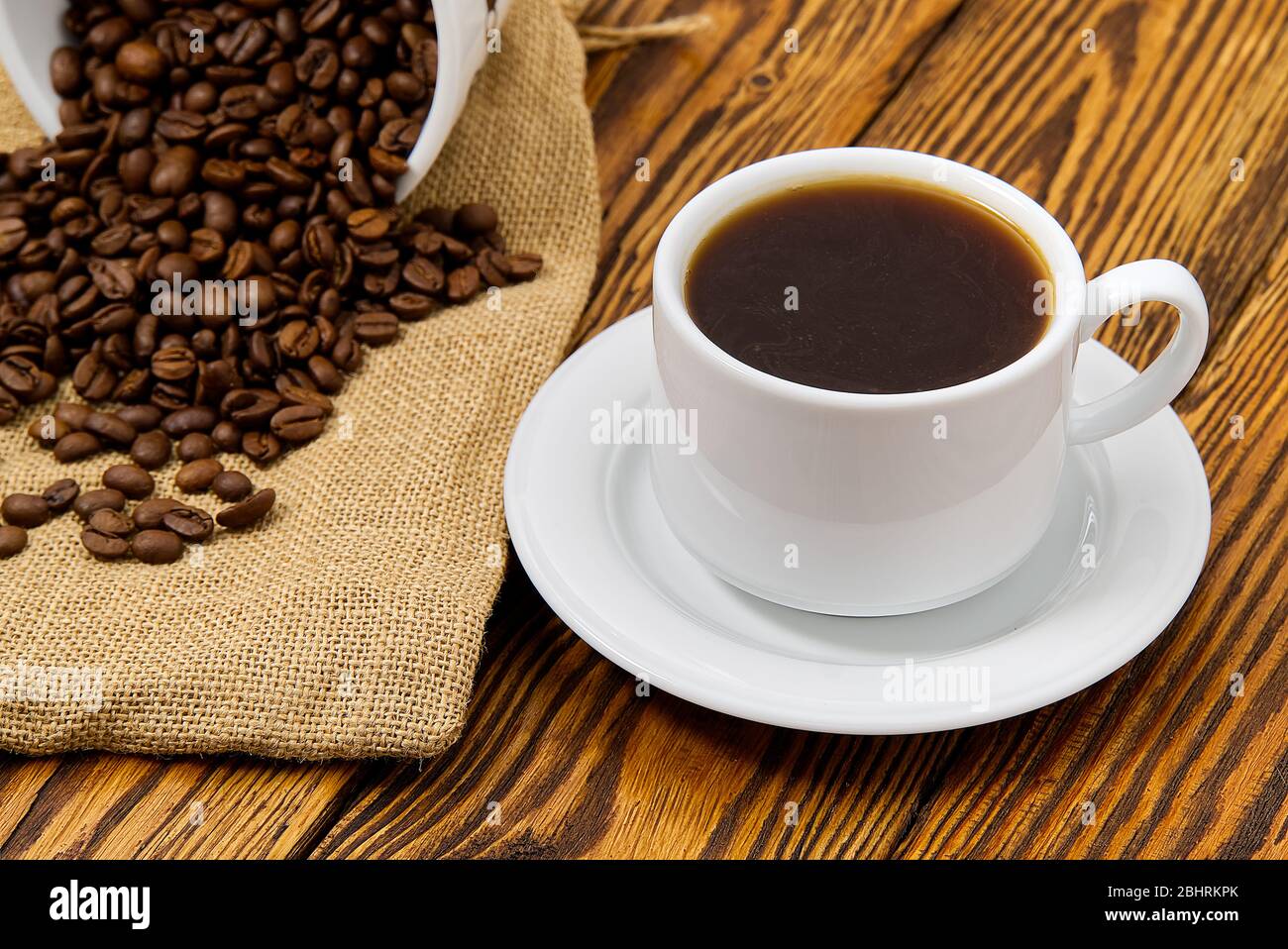 Weiße Kaffeetasse mit aromatischem Kaffeegetränk auf braunem Holztisch. Kaffeesamen auf antikem Holztisch mit Tasse Kaffee Getränk Stockfoto