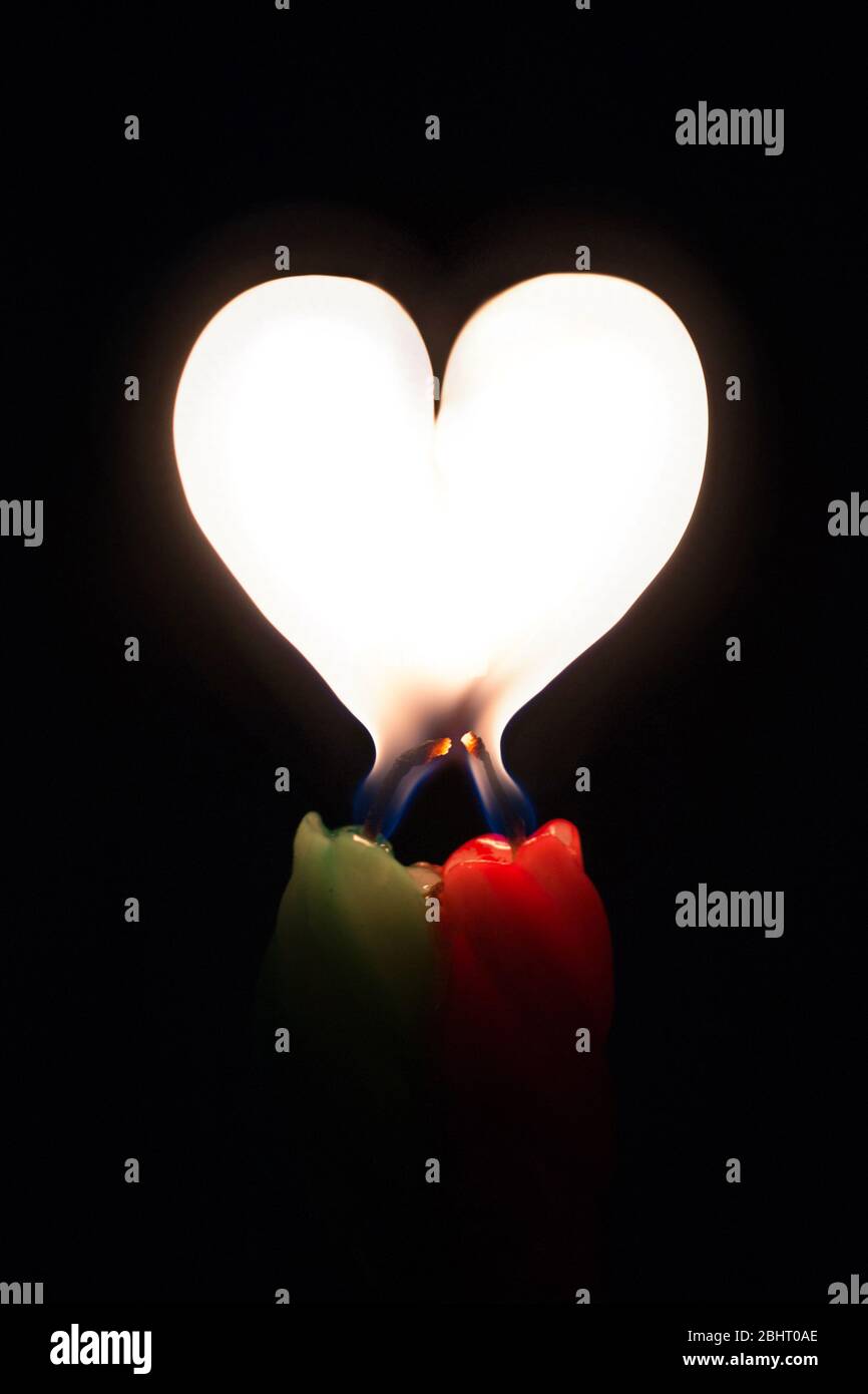 Zwei farbige brennende Kerzen mit einer Flamme in Form eines Herzens auf schwarzem Hintergrund Stockfoto