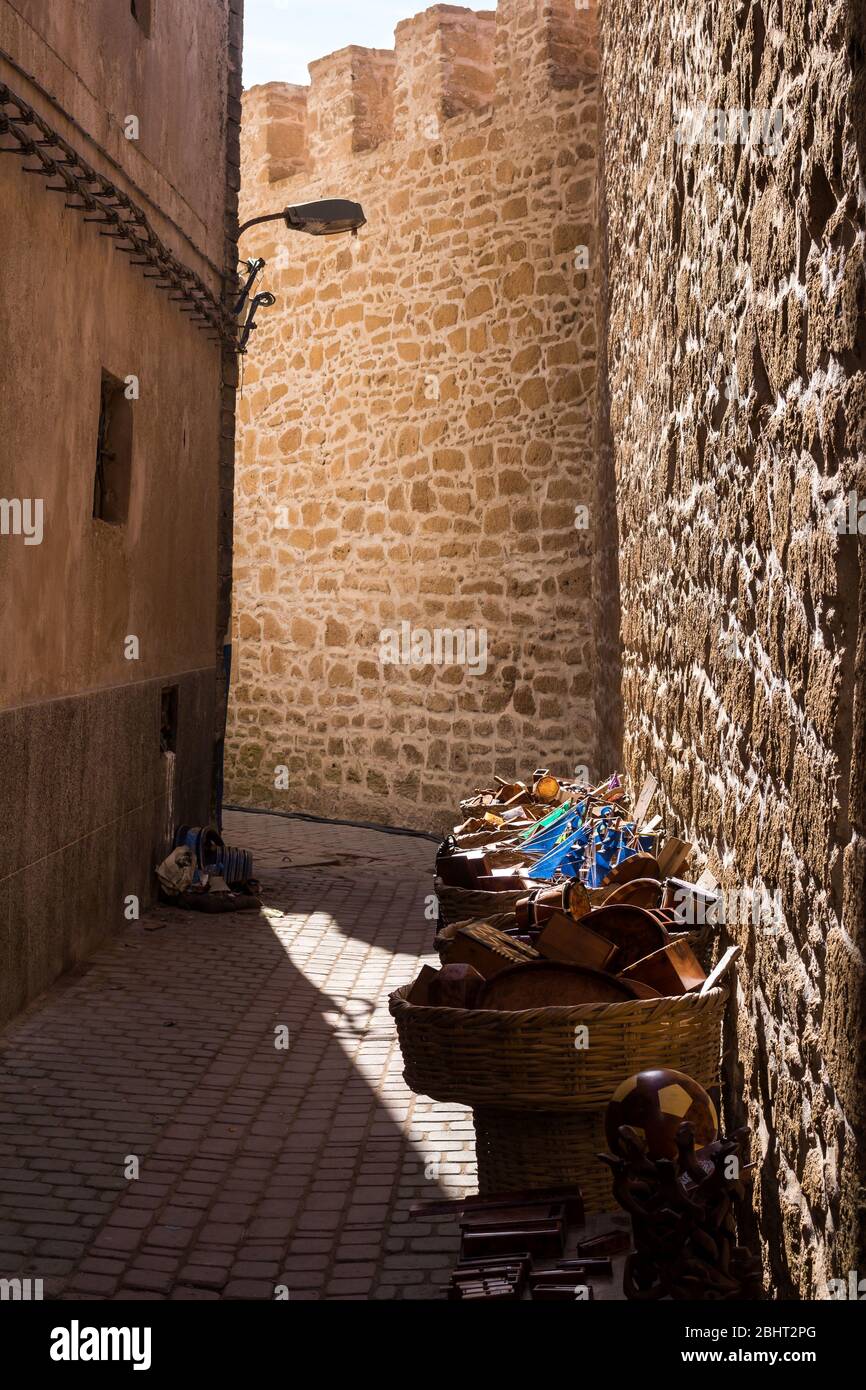 Straße, die die Steinmauer säumen, teilweise von der Sonne beleuchtet. Körbe mit ausgestellten Waren eines Ladens. Essaouira, Marokko. Stockfoto