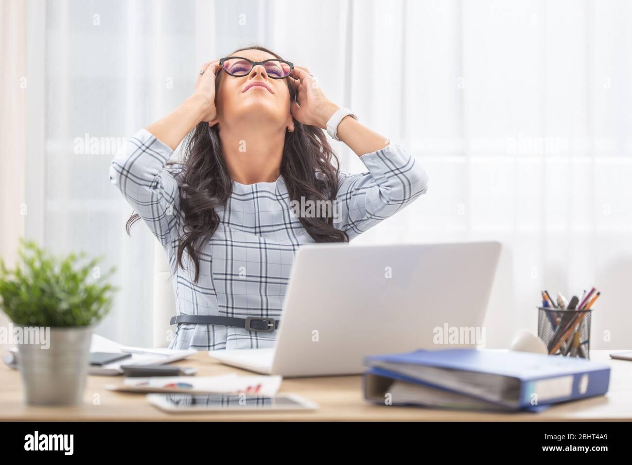 Büroassistentin, die sich auf ihrem Stuhl zurücklehnt und den Kopf in den Händen hält, da sie bei der Arbeit Kopfschmerzen hatte. Stockfoto