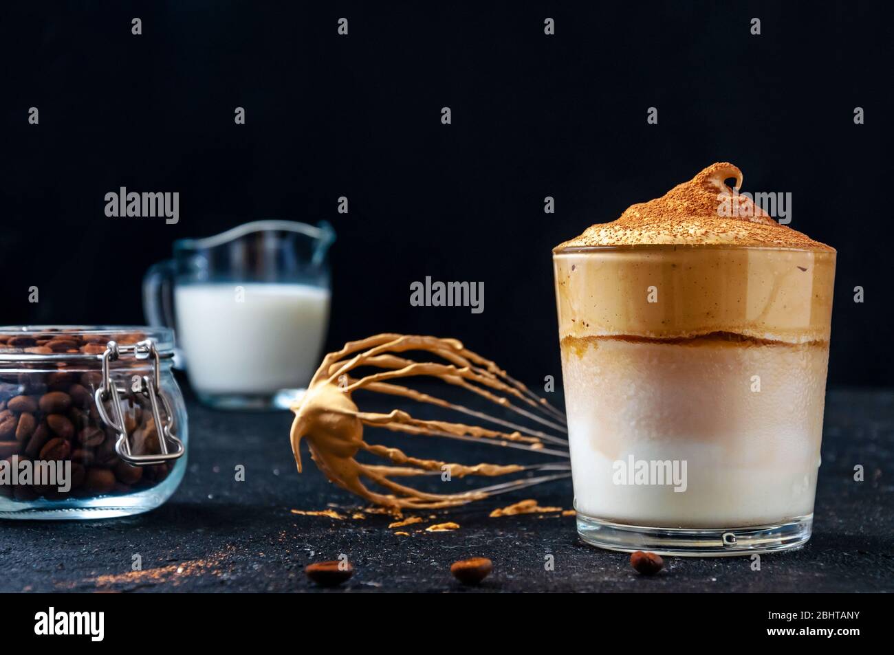 Iced Dalgona Kaffee mit Corolla, Milch und Kaffeebohnen auf dunklem Hintergrund. Trendy Cremigen Schlagsahne Kaffee. Südkoreanischer Kaltsommer-Drink. Stockfoto