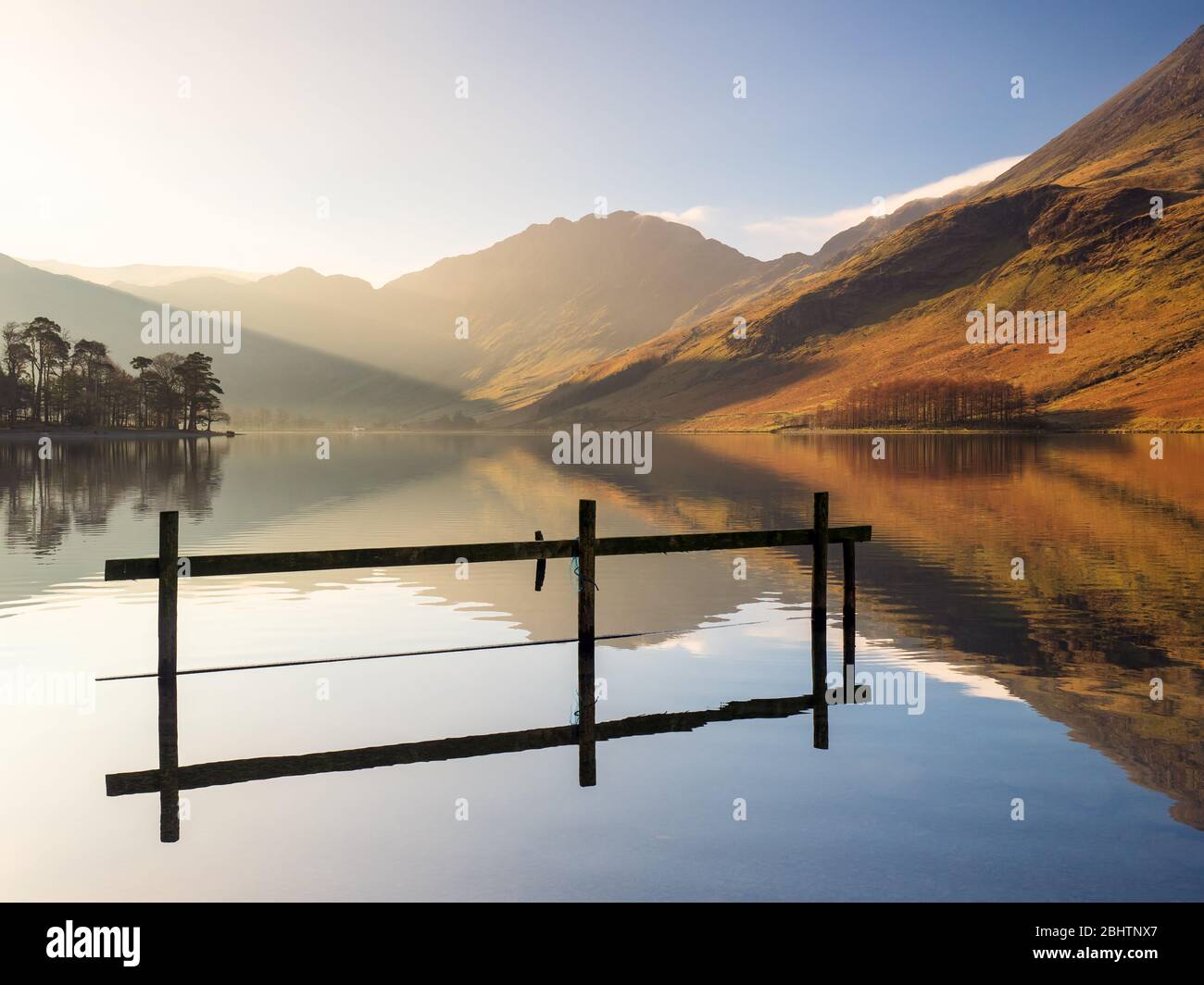 Ein klarer Frühlingsmorgen in Buttermere im Lake District. Das ruhige Wasser des Sees erzeugt perfekte Reflexionen des Zauns, der Bäume und der Berge. Stockfoto