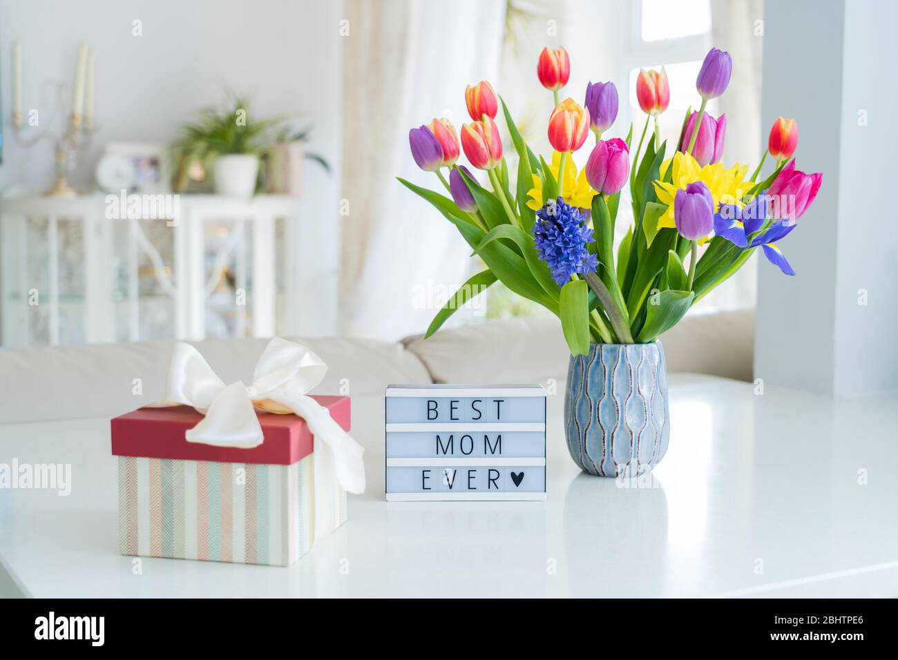 Alles gute zum Muttertag Hintergrund. Bunte Frühlingsblumen Bouquet in Vase, Geschenkbox mit Satinband und Leuchtkasten mit Worten Best mom ever auf weißem Marmor Stockfoto