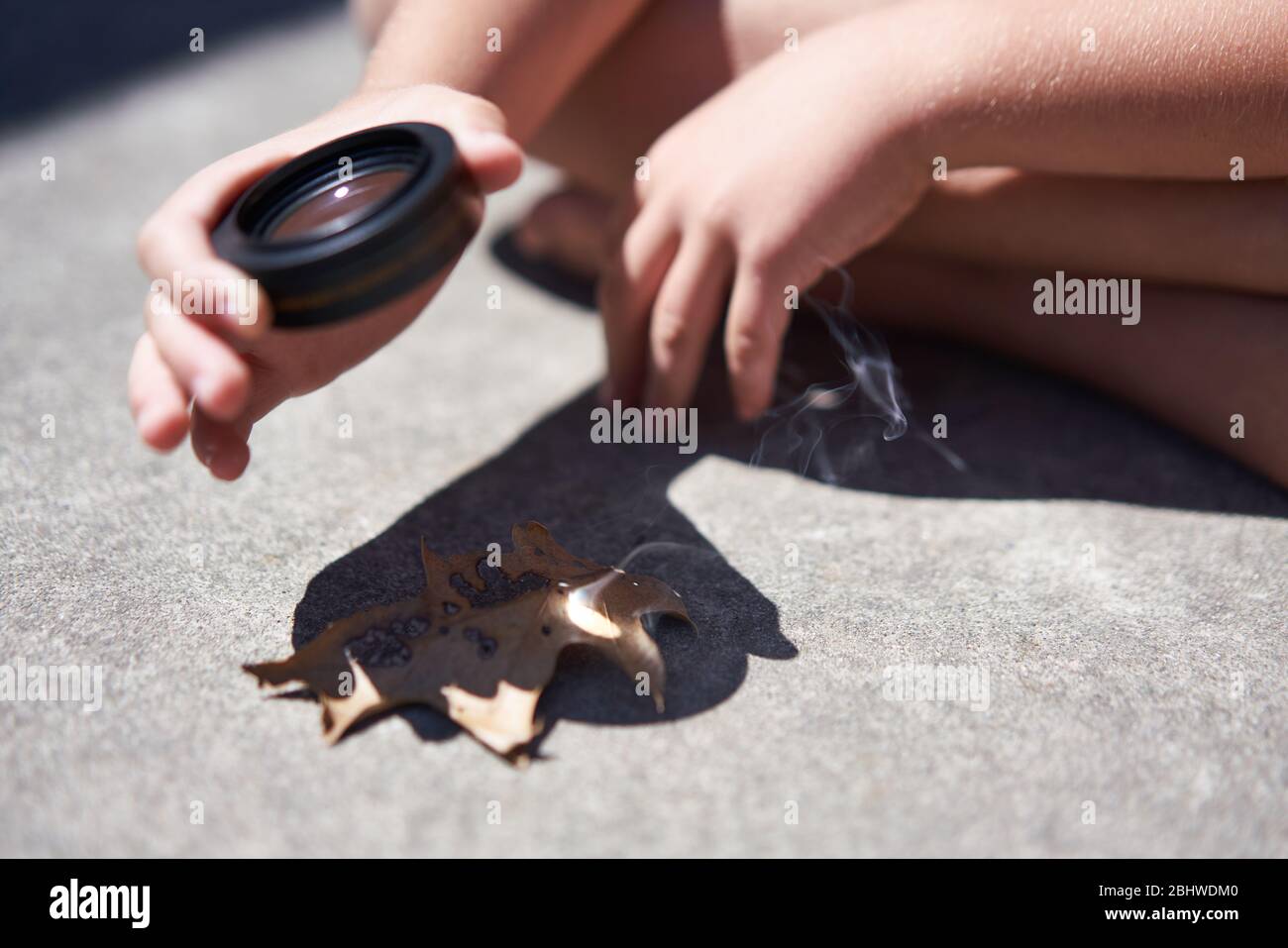 Hände und Finger halten eine konvexe Linse und fokussieren Sonnenlicht auf Blätter, die sie entzünden, rauchen und brennen. Stockfoto