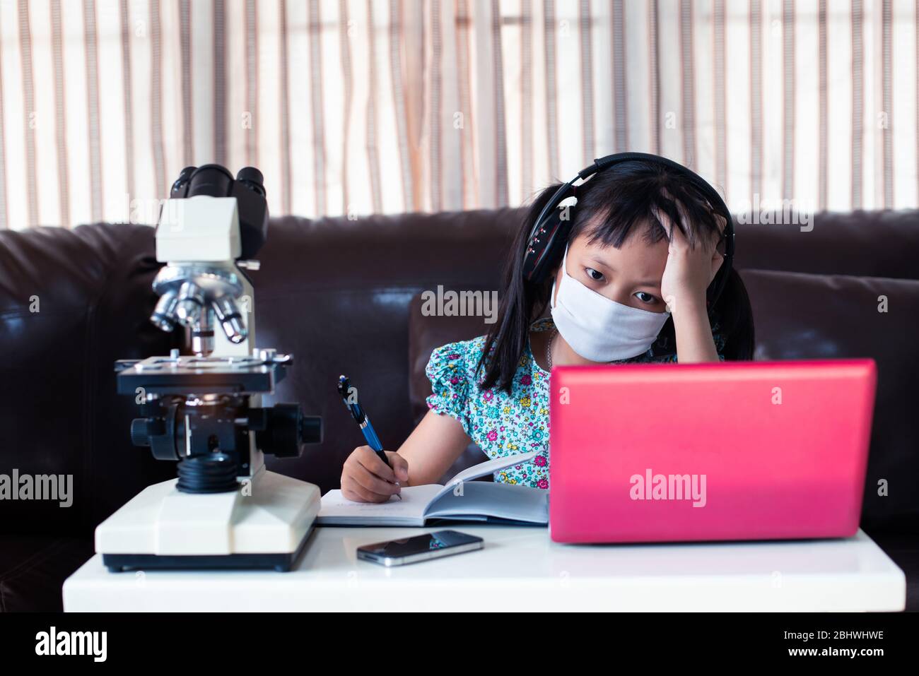 Stress Kind Mädchen tragen Gesichtsmaske und Kopfhörer online lernen, indem sie Laptop und Mikroskop zu Hause, Fernunterricht Stockfoto