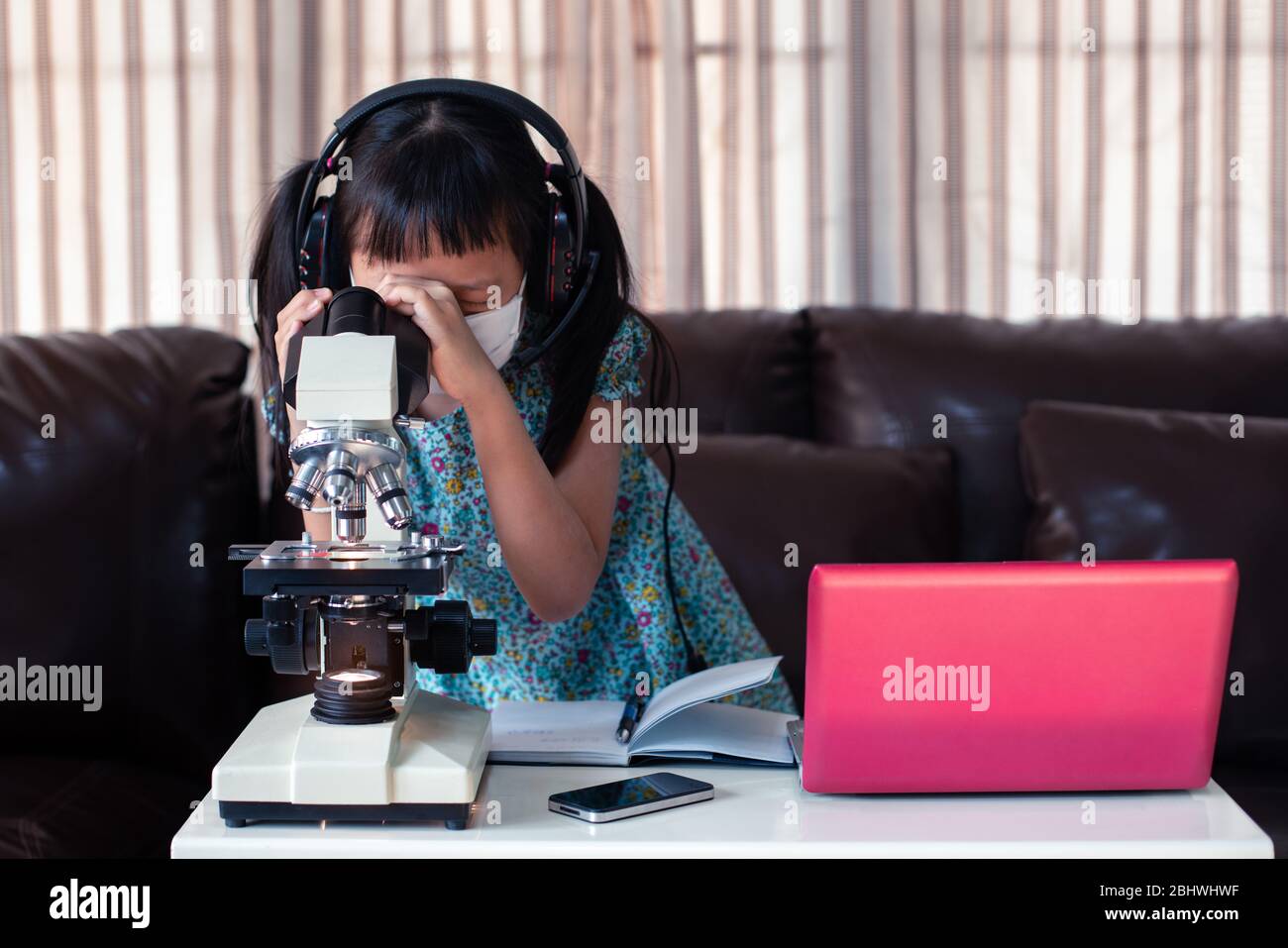 Kleines Kind Mädchen tragen Gesichtsmaske und Kopfhörer online lernen, indem sie Laptop und Mikroskop zu Hause, Fernunterricht. Stockfoto