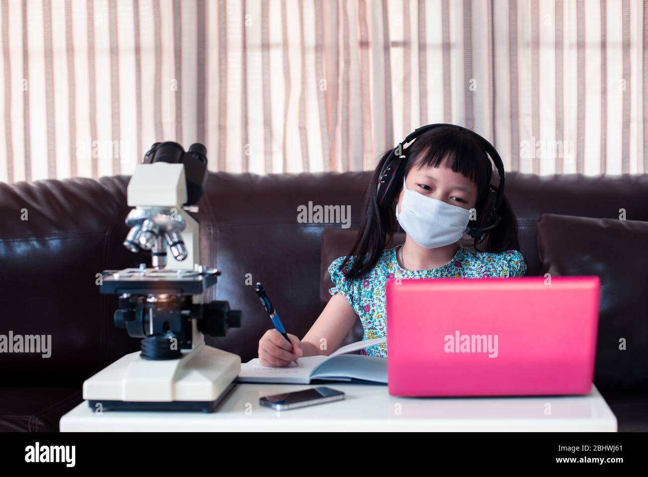 Kleines Kind Mädchen tragen Gesichtsmaske und Kopfhörer online lernen, indem sie Laptop und Mikroskop zu Hause, Fernunterricht. Stockfoto