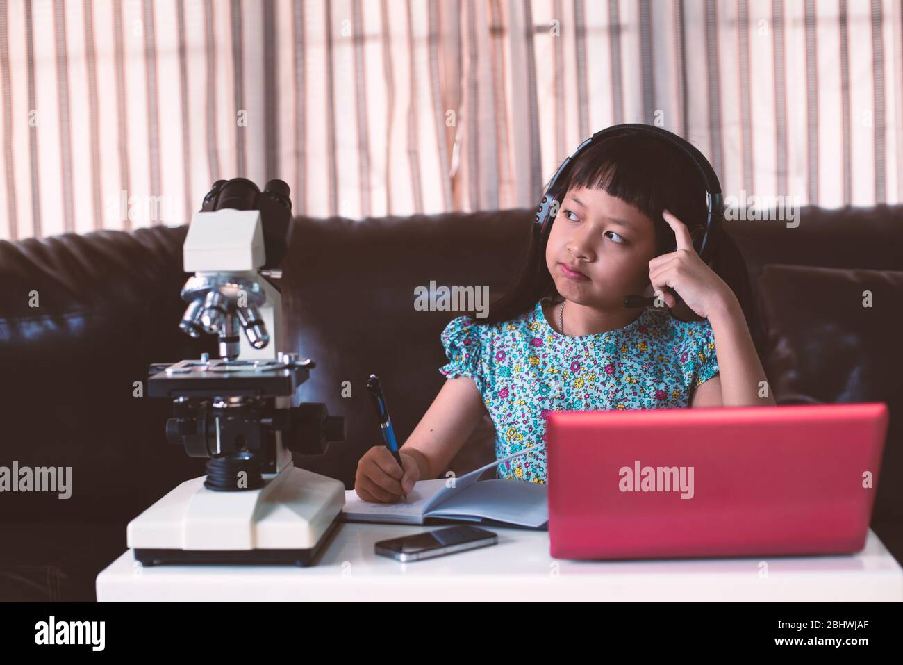 Asiatische kleines Kind Mädchen denken und tragen Kopfhörer online lernen, indem sie Laptop mit Mikroskop zu Hause, Fernunterricht Stockfoto