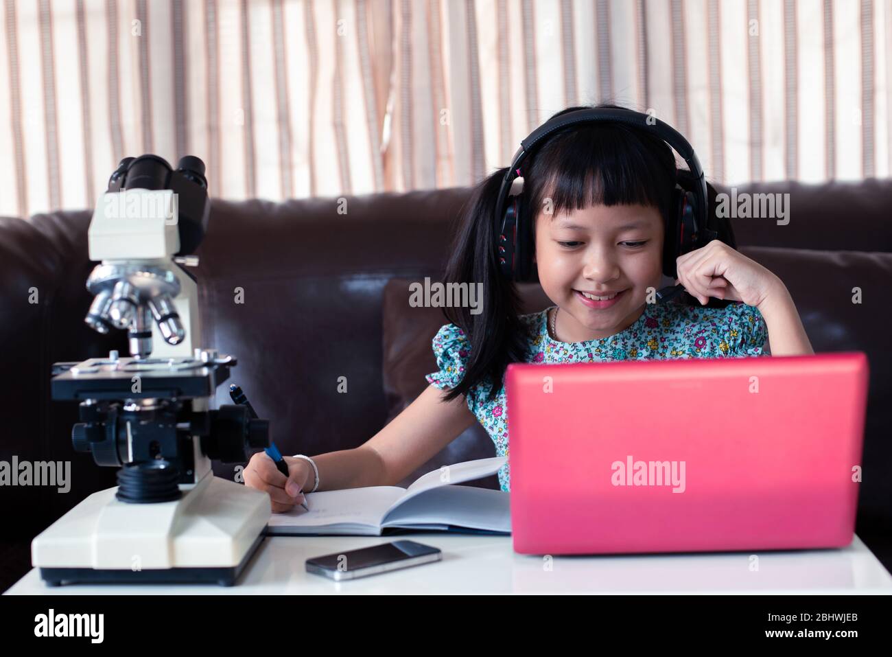 Asiatische kleines Kind Mädchen tragen Kopfhörer online lernen, indem sie Laptop und Mikroskop zu Hause, Fernunterricht Stockfoto