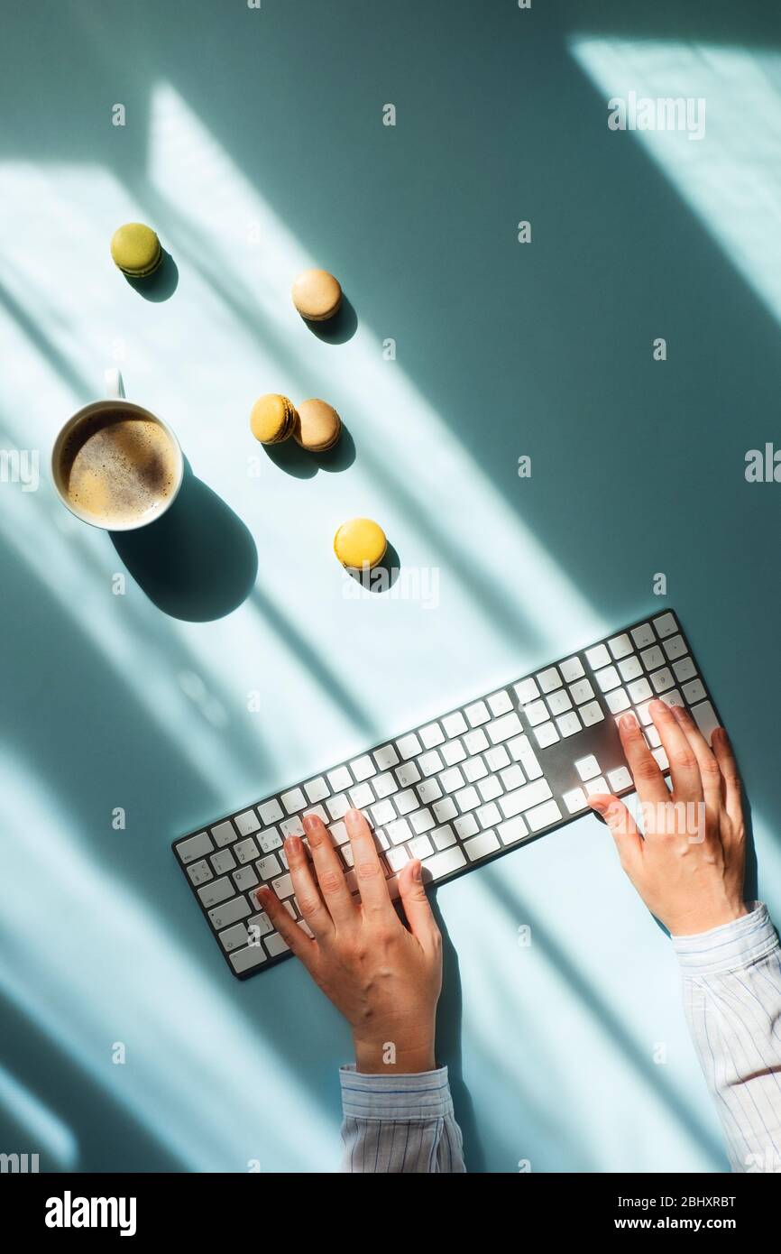Arbeitsplatz zu Hause am Fenster. Flacher Lat mit einer Tasse Kaffee und Makronen im Sonnenlicht und Frauen geben die Hände auf der Tastatur. Stockfoto