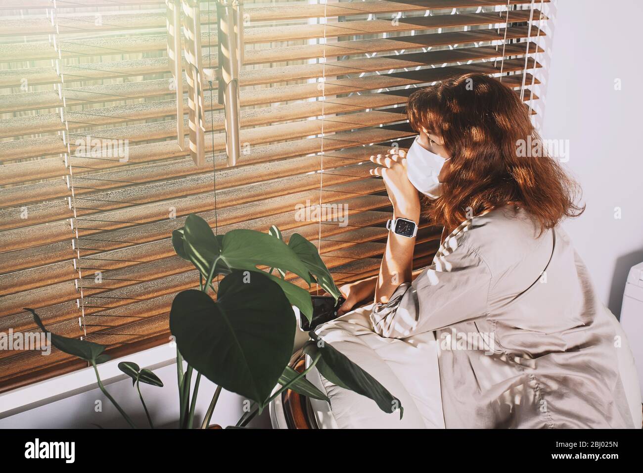Frau mittleren Alters, die eine Gesichtsmaske trägt und von Jalousien ausguckt. Frau in Quarantäne schaut durch das Fenster. Corona Covid-19 Virenschutz. Stockfoto