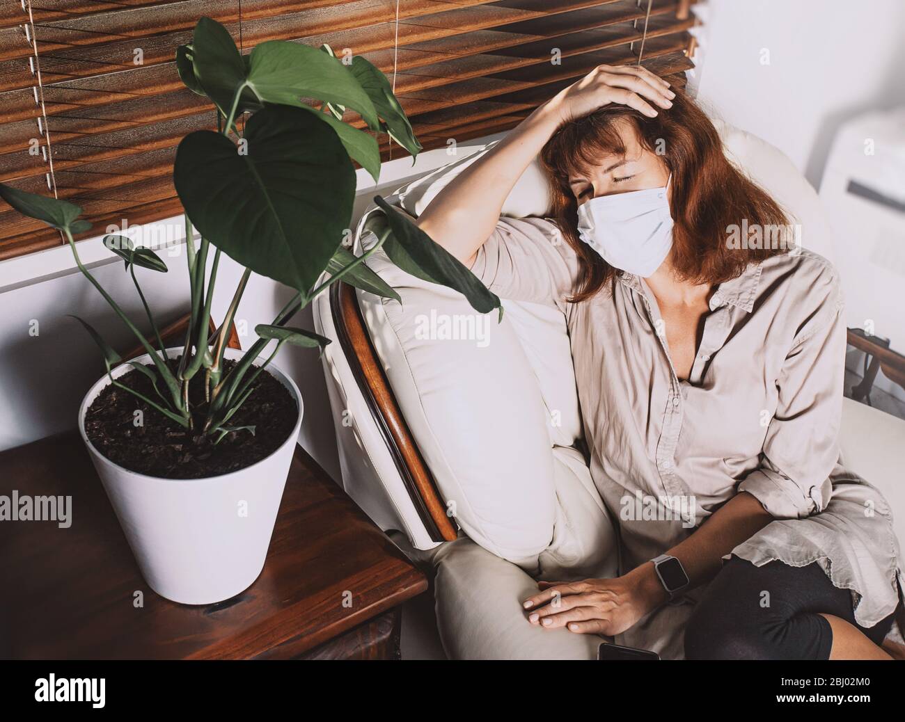 Frau mittleren Alters trägt eine Gesichtsmaske, sitzt auf einem Sessel mit geschlossenen Augen, fühlt sich erschöpft oder krank. Sperrung der Corona COVID-19-Quarantäne. Stockfoto