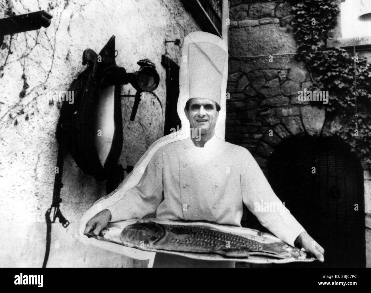 Küchenchef Paul Bocuse posiert mit einem riesigen Fisch auf einer Platte in einem rustikalen Innenhof Stockfoto