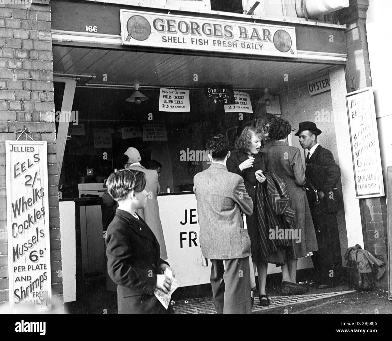 George's Bar - Shell Fish Fresh Daily - EINE Schlange von Leuten wartet darauf, bedient zu werden, aber eine Frau, die gerade ihre Bestellung erhalten hat, scheint nicht allzu beeindruckt vom Geschmack. - 1950er Jahre Stockfoto
