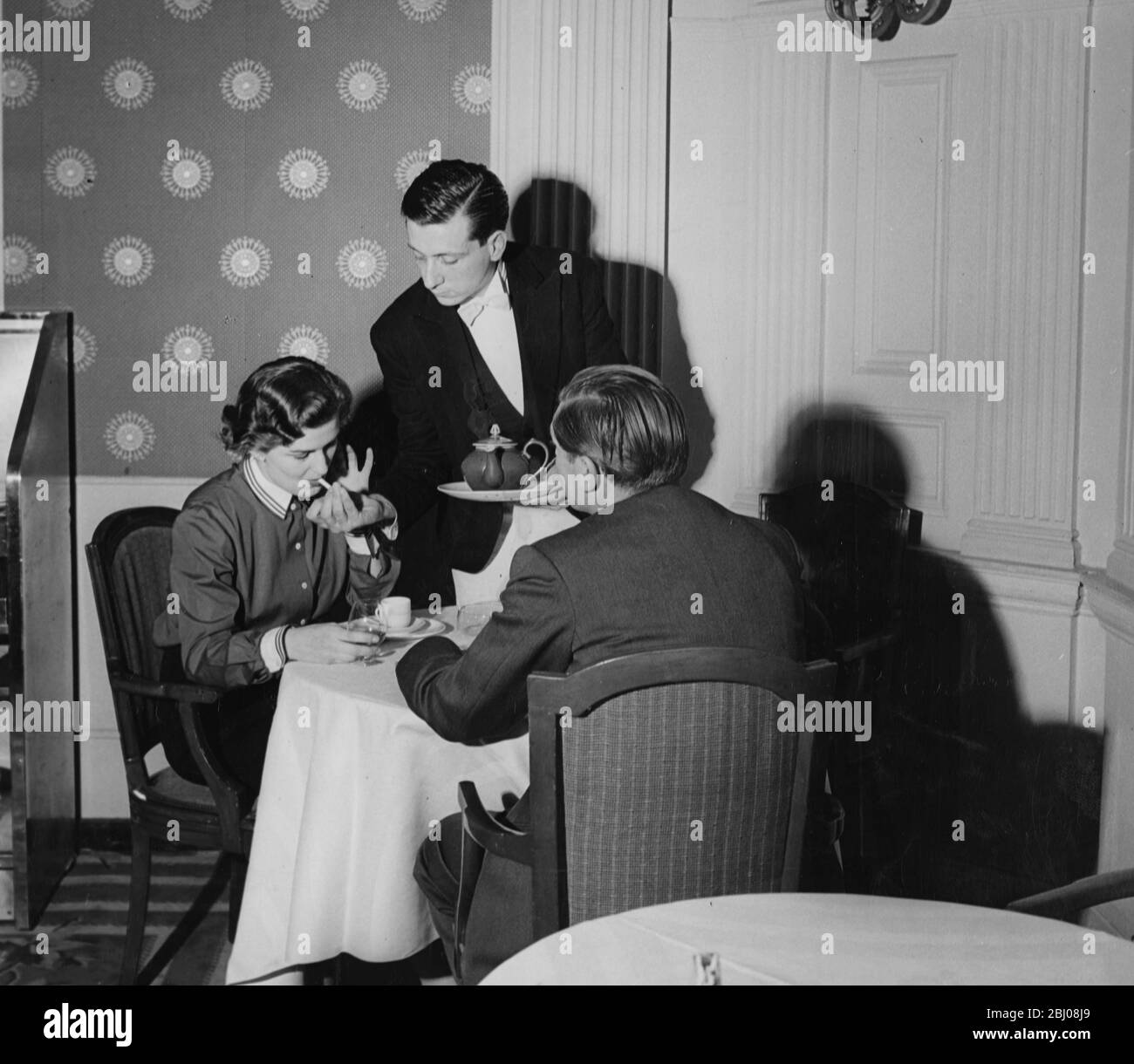 Natale Rusconi kehrt zu seinem Tagesjob als Kellner zurück, während er ein paar Tee serviert und einer Dame eine Zigarette anzündet. Die letzten zehn Tage wurde er als Dolmetscher für die italienischen Filmstars rekrutiert, die für das Filmfestival, England - 20. November 1954 nach London strömten Stockfoto