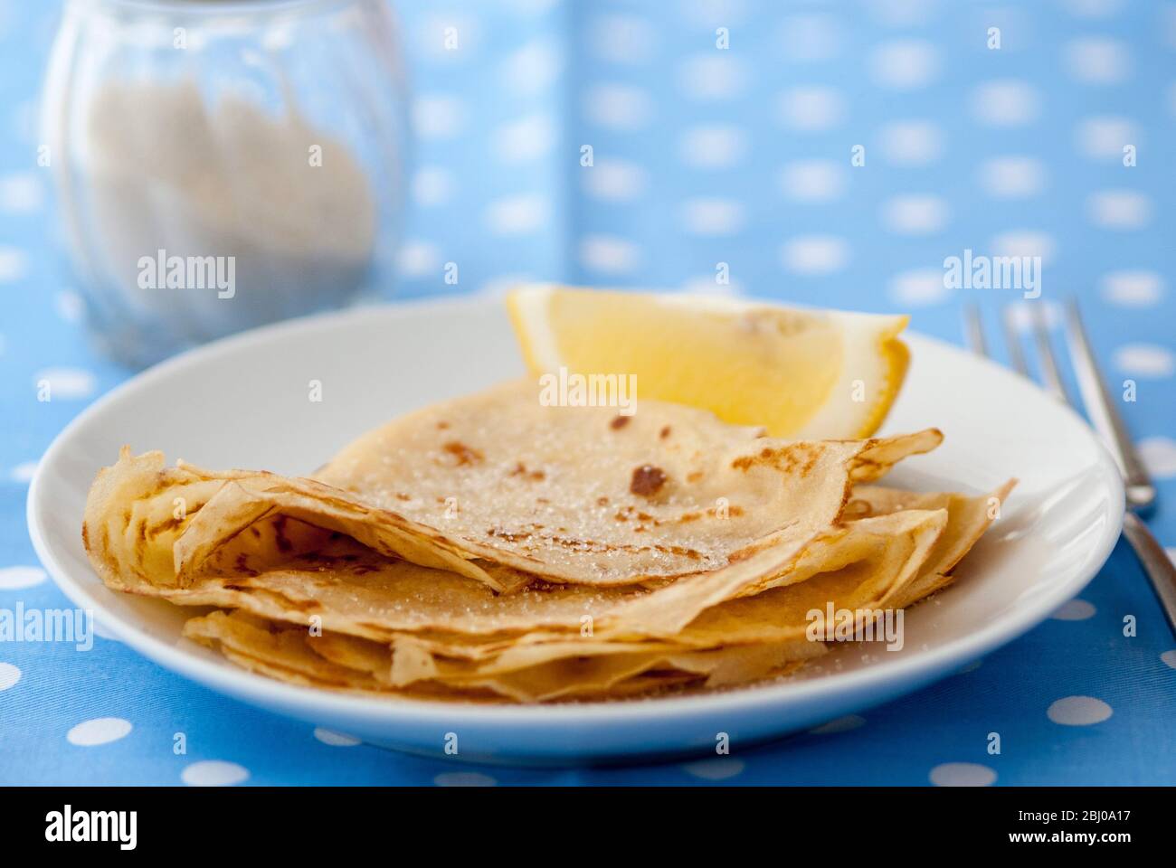 Klassische Pfannkuchen in vier gefaltet und auf weißem Teller mit Zitronenkeil und Zucker auf blau und weiß gefleckt Hintergrund gestapelt Stockfoto