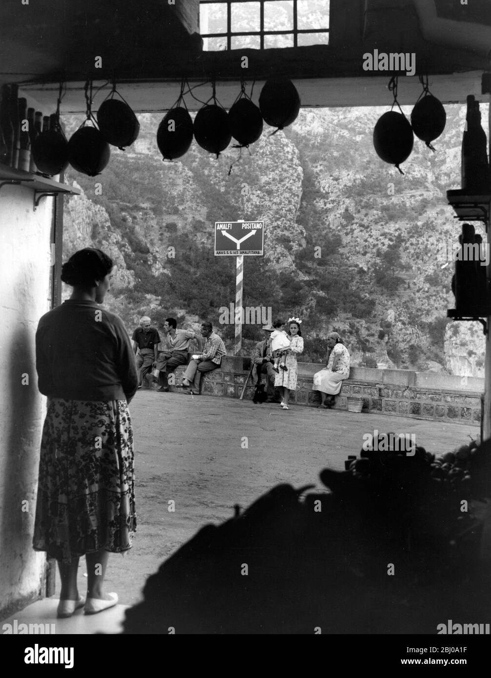 Eine Frau Ladenbesitzerin blickt auf die Straße, wo Einheimische plaudern und eine junge Frau ein Kleinkind hält. Ordentlich Positano, Italien Stockfoto