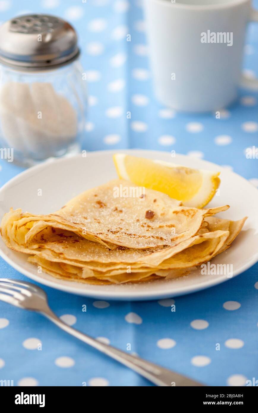 Klassische Pfannkuchen in vier gefaltet und auf weißem Teller mit Zitronenkeil und Zucker auf blau und weiß gefleckt Hintergrund gestapelt Stockfoto