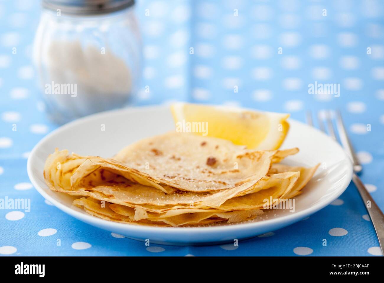 Klassische Pfannkuchen in vier gefaltet und auf weißem Teller mit Zitronenkeil und Zucker auf blau und weiß gefleckt Hintergrund gestapelt Stockfoto