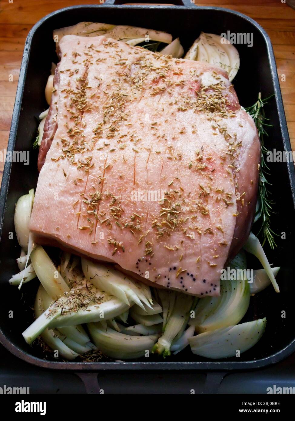 Ein Stück Schweinebauch, das mit Fenchelsamen auf dem Bett der Zwiebeln und des fenchels in der Metallröstung gebraten werden kann - Stockfoto