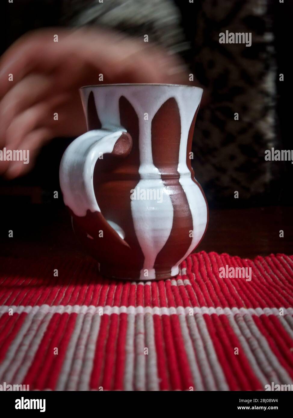 Eine Tasse Kaffee oder Tee in einem streifenbedeckten Töpferbecher auf einer gestreiften Tischdecke abholen - Stockfoto