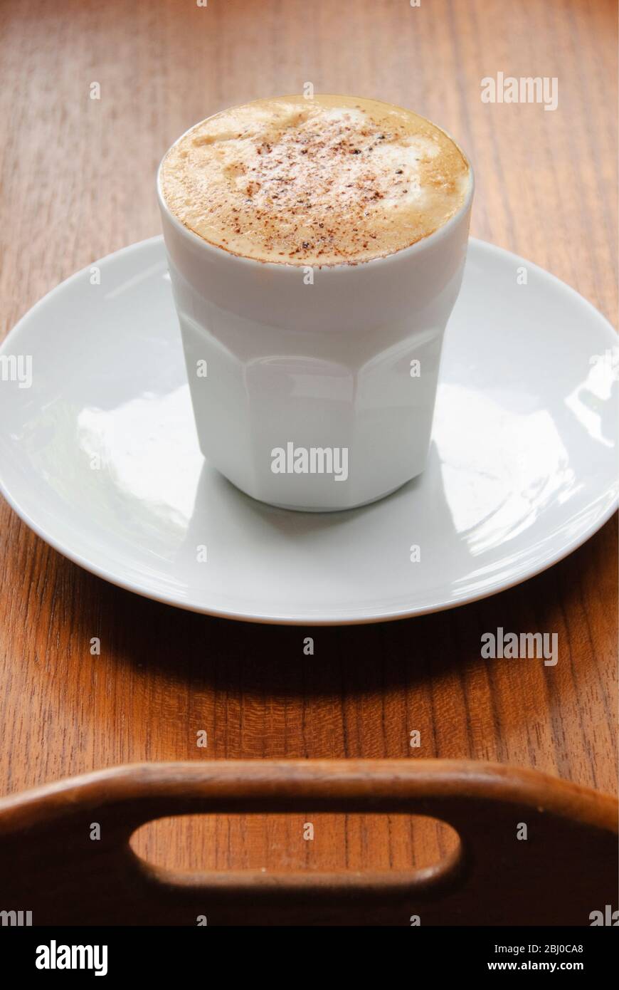 Cappuccino mit schaumiger Spitze in weißer, griffloser Tasse auf braunem Teakholztablett - Stockfoto