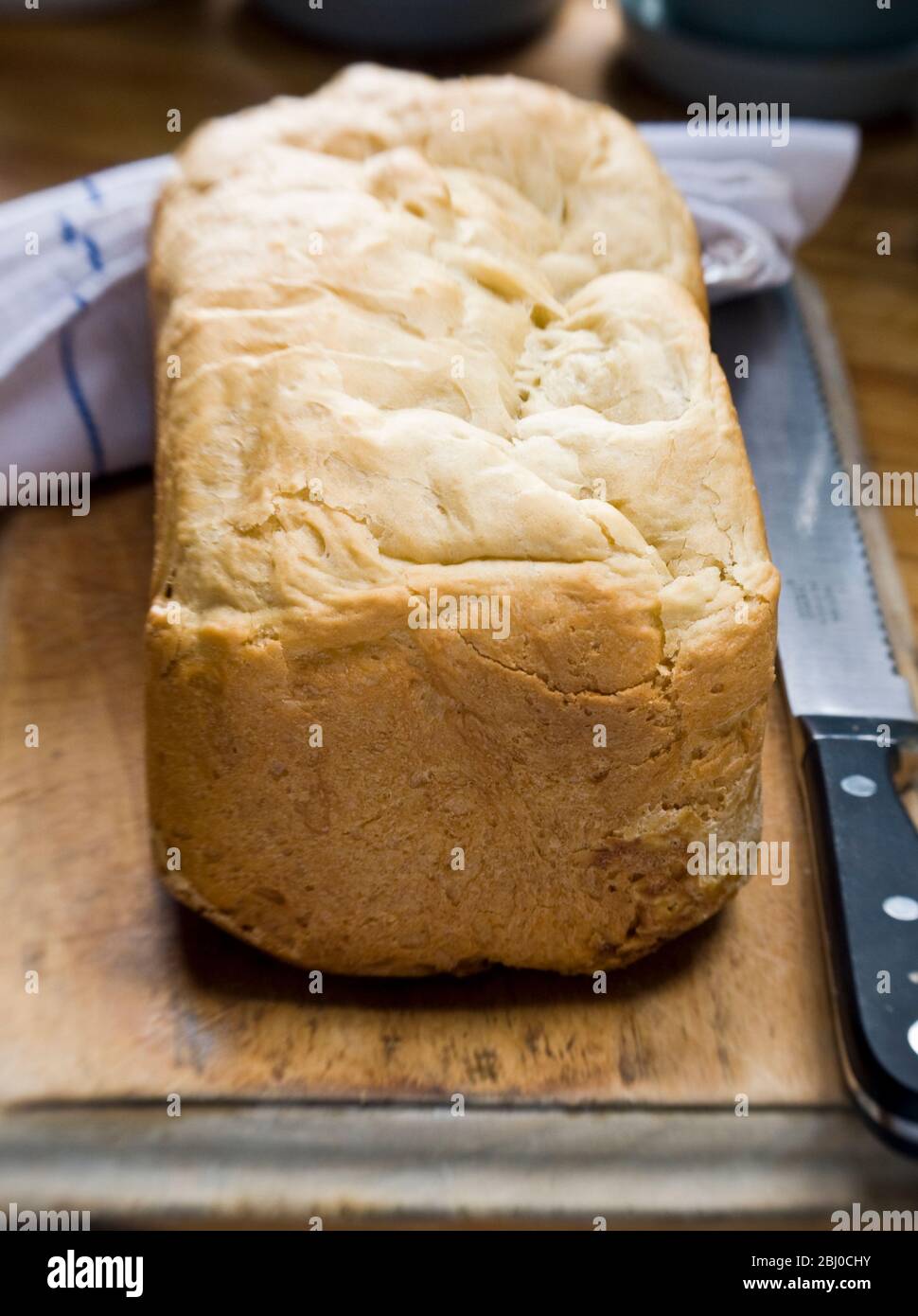 Weißer knuspriger Laib mit Bio-Mehl hergestellt in Elektro-Brotbackautomat auf alten Holz Brottafel mit gezackten Messer - Stockfoto