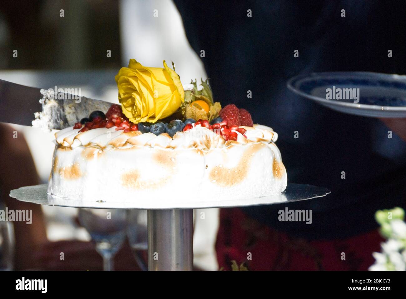 Festtorte aus leichter Zitrone und Passionsfrucht Mousse mit weicher Meringue-Topping, frischen exotischen Früchten und gelben Rosen, die im Sommer draußen geschnitten werden Stockfoto