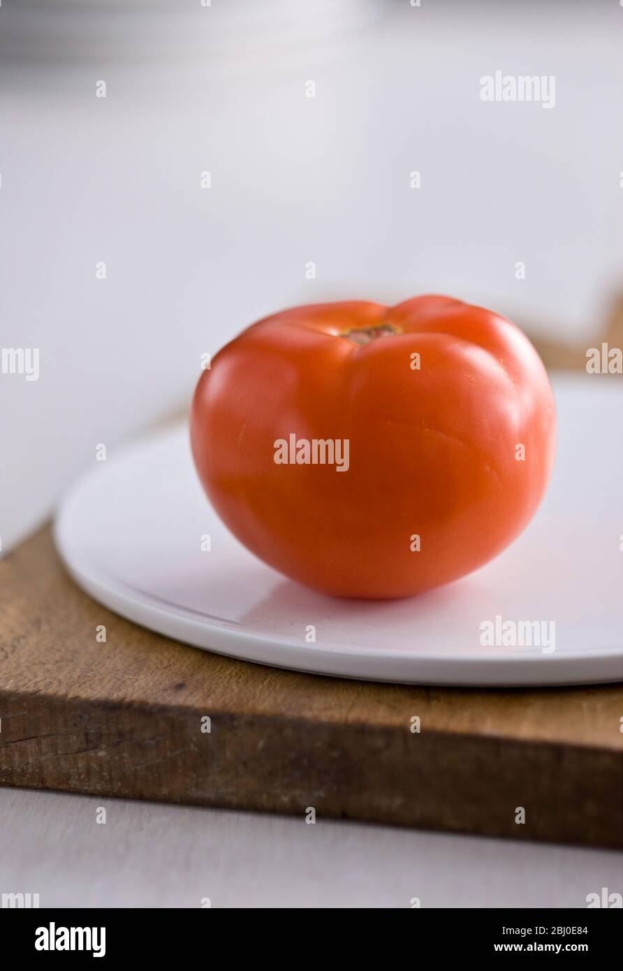 Einzelne rote Tomate auf weißer Platte auf Holzbrett - Stockfoto