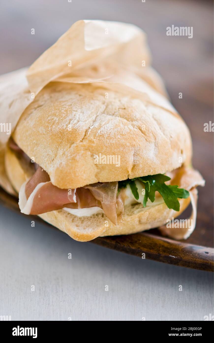 Italienisches Schinkenbrot, parmaschinken, Mozzarella-Käse und frische Rucola auf leichtem Ciabatta-Brot, in fettabweisem Papier verpackt. - Stockfoto