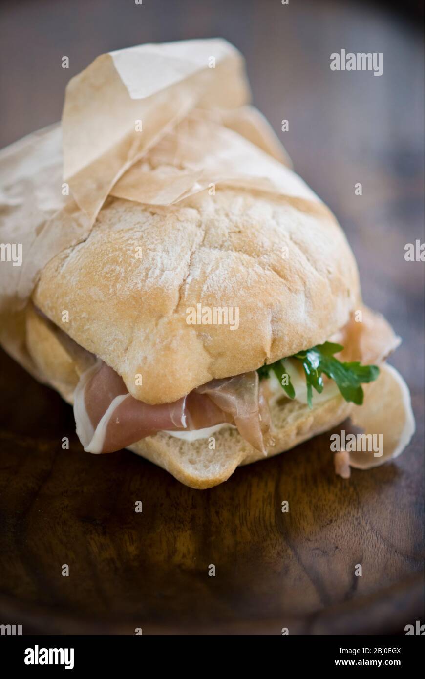 Italienische Schinken Sandwich, parma Schinken, Mozzarella Käse, und frech Rakete auf Licht Ciabatta Art Brot, in fettfreien Papier verpackt. - Stockfoto
